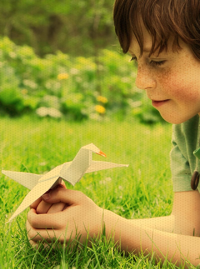 Little boy laying in grass, smiling and holding an origami bird made of paper